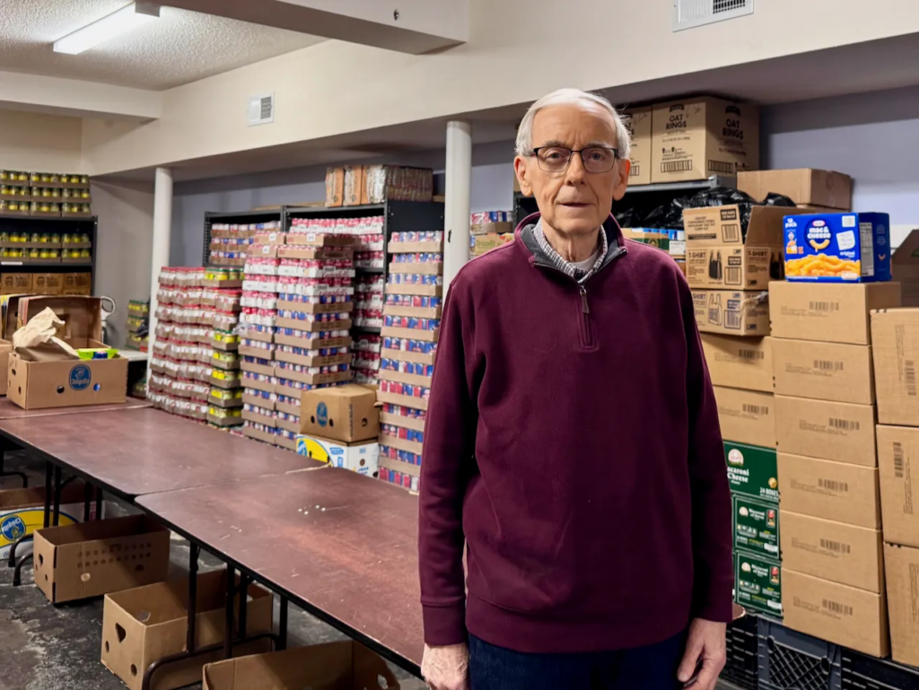 Older man with maroon sweater standing in front of boxes of nonperishable food.